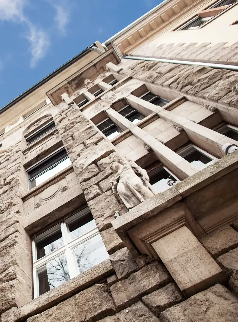 The outside facade of an old beige building looking from the bottom up towards the top where there is a corner of blue sky.