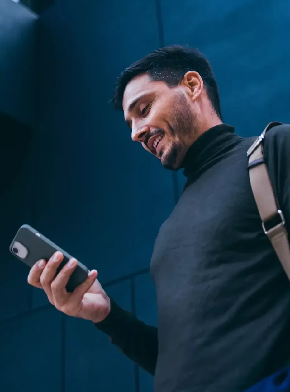 A man wearing a black turleneck is looking down at his smartphone which he is holding in his right hand. He has a bag on his left shoulder and he is smiling.